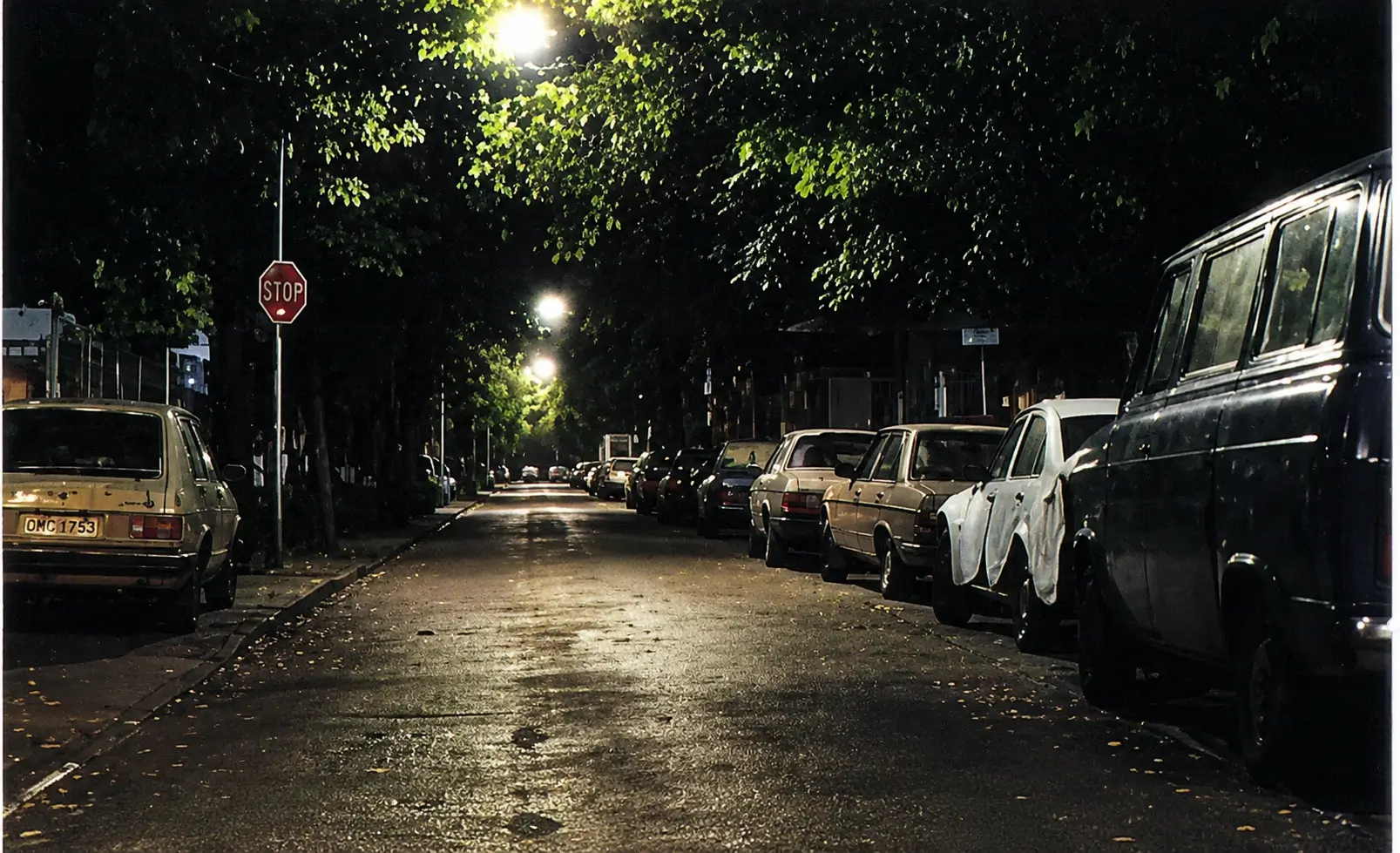 unevenly lit street with cars parked on the right side shoulder