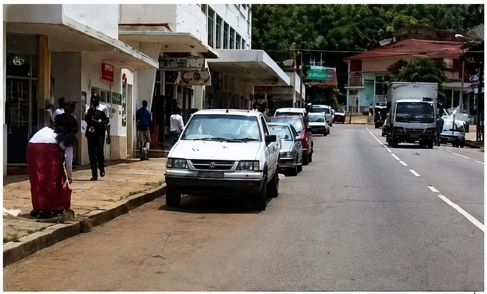 driver's view of a built up area with oncoming traffic and cars parked on the left shoulder